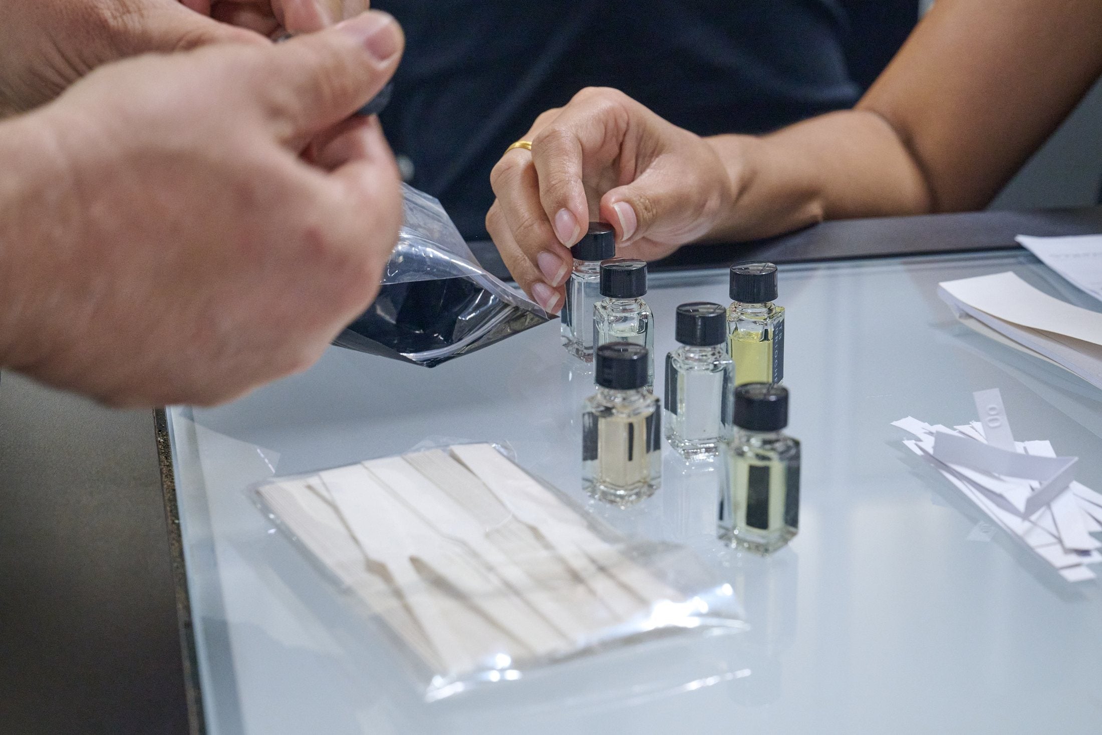 Hands compare fragrance blotters and small perfume bottles across a workshop table.